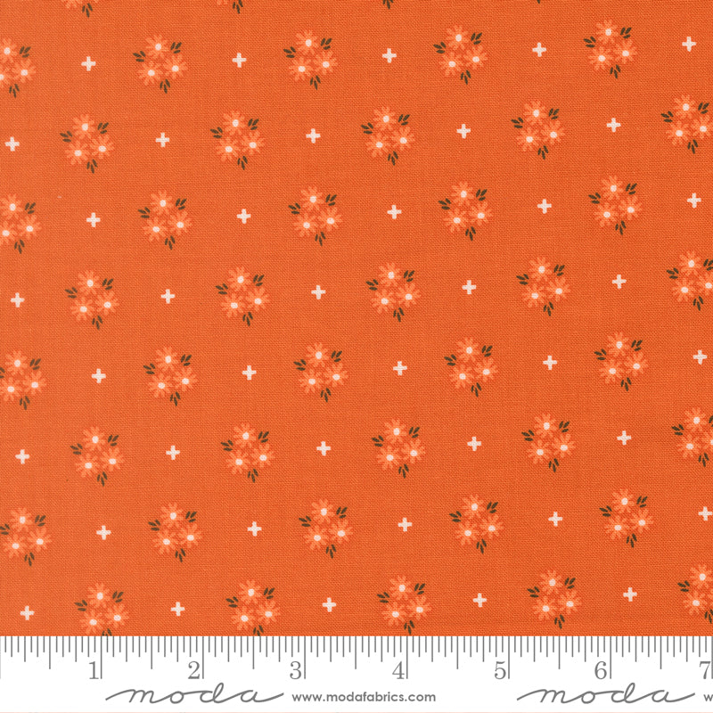 Fabric swatch with an orange background and lighter orange and white flowers with white plus signs scattered around. Ruler at bottom for scale.