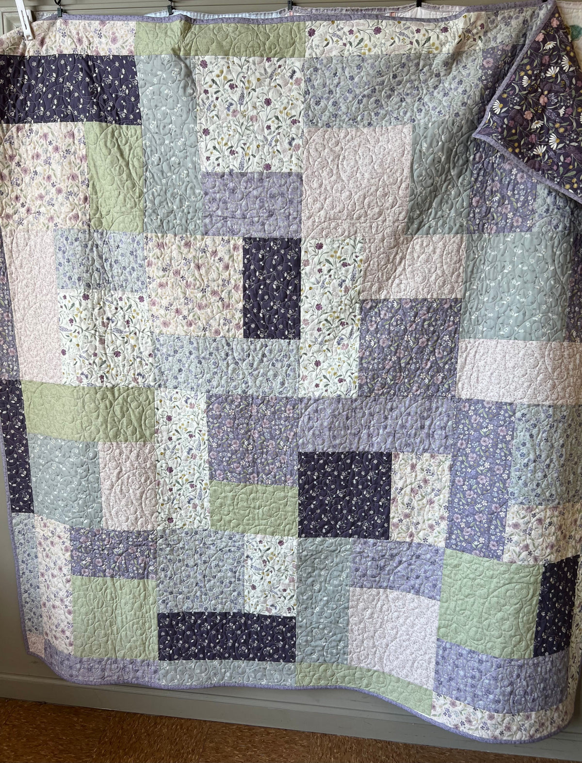 A patchwork quilt hanging flat against a wall, featuring a mix of solid and floral patterned fabric squares in muted lavender, sage green, cream, and dark purple tones.