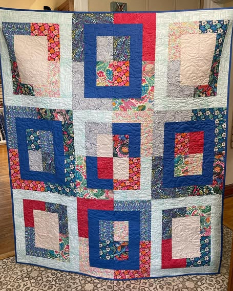 A handmade patchwork quilt hanging flat in a home setting, featuring a rectangle style design with blue borders, red and white centers, and various floral patterned fabrics.