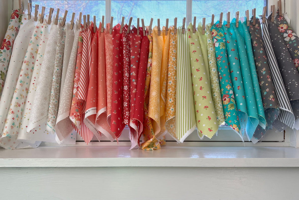 A collection of colorful patterned fabric bolts hanging from wooden clothespins on a window sill, arranged in a rainbow-like gradient of red, orange, yellow, green, blue, and gray.