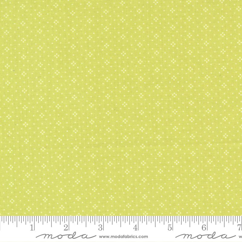 A close-up of a fabric pattern featuring a light yellow-green background with a repeating small white floral or starburst motif. A white ruler along the bottom edge shows measurement markings and the Moda Fabrics brand name.