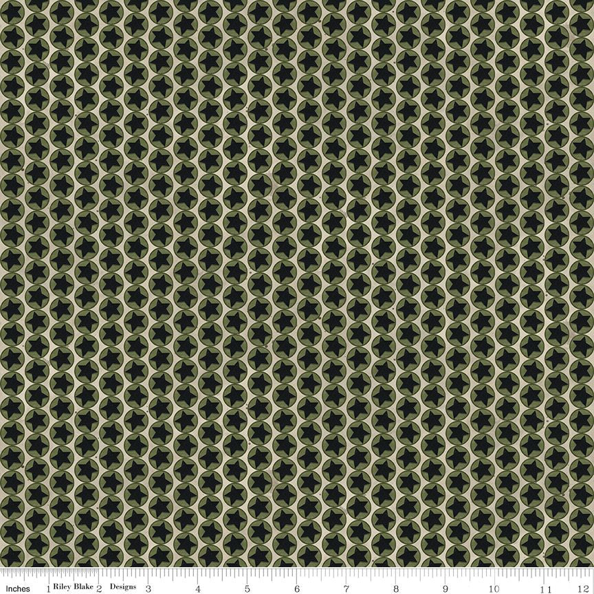 A close-up of a repeating fabric pattern with dark olive green and black geometric starbursts inside interlocking hexagons, with a ruler along the bottom edge showing scale.