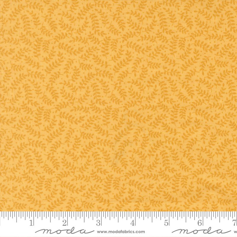 A close-up of a fabric pattern showing a warm mustard yellow background with a repeating small leafy vine motif in a slightly darker golden yellow. A white ruler with black markings is visible along the bottom edge, and the Moda Fabrics logo and website are printed at the bottom.