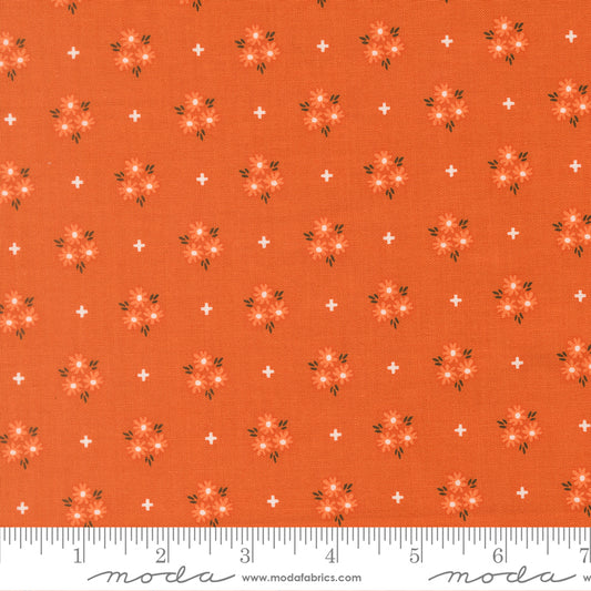 Fabric swatch with an orange background and lighter orange and white flowers with white plus signs scattered around.  Ruler at bottom for scale.