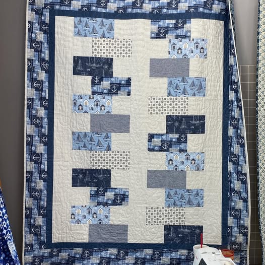 A handmade patchwork quilt featuring a nautical theme with blue, navy, and white fabrics. The quilt has a central cream-colored block pattern surrounded by blue and navy borders with printed anchor and ship designs.