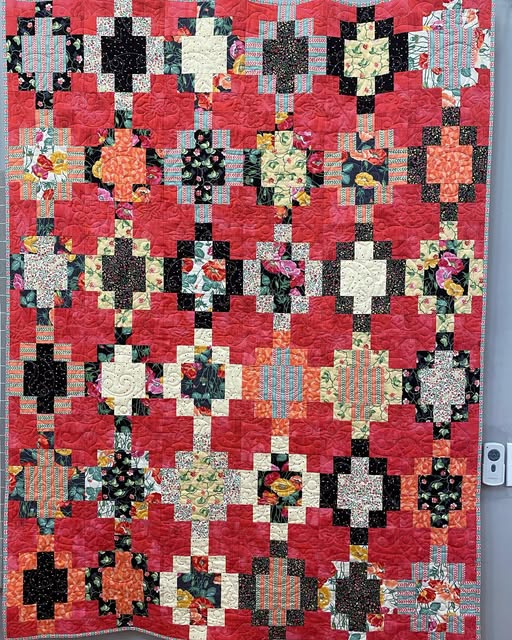 A handmade quilted small-scale quilt featuring a repeating geometric cross pattern in vibrant red, black, cream, and various floral fabrics, shown hanging flat against a gray wall.