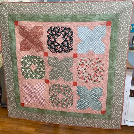 A handmade patchwork quilt hanging flat on a display stand in a room. The quilt features a central grid of nine floral and geometric blocks in muted pink, sage green, and black, surrounded by a wide sage green border and a light beige floral outer edge.