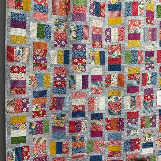 A colorful patchwork quilt hanging on a wall, made of many small fabric squares in floral, striped, and solid prints on a light gray background.