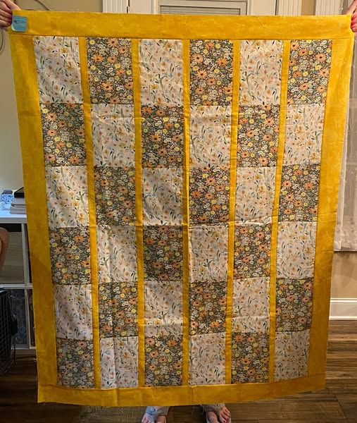 A handmade patchwork quilt held up by a person in a home setting. The quilt features a wide mustard yellow border and alternating vertical stripes of solid mustard yellow and floral patterned panels.