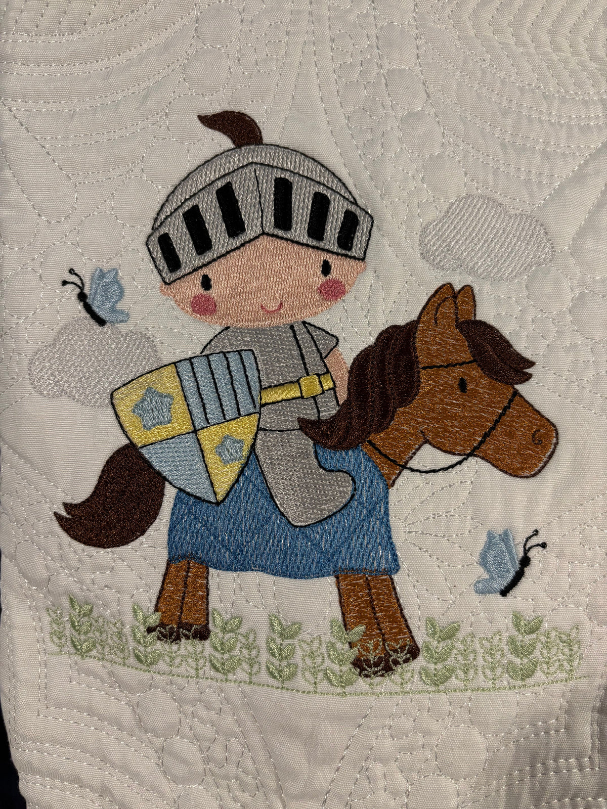 Close-up of a quilted fabric featuring an embroidered knight in gray armor and a blue tunic riding a brown horse, with small blue butterflies and green leafy stems on a white quilted background.