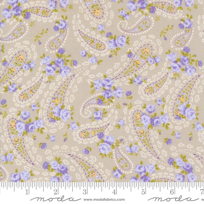 A close-up of a fabric pattern featuring lavender and purple flowers, green leaves, and cream paisley motifs on a warm beige background, with a measuring ruler along the bottom edge showing scale. The brand name 'moda' is visible at the bottom.