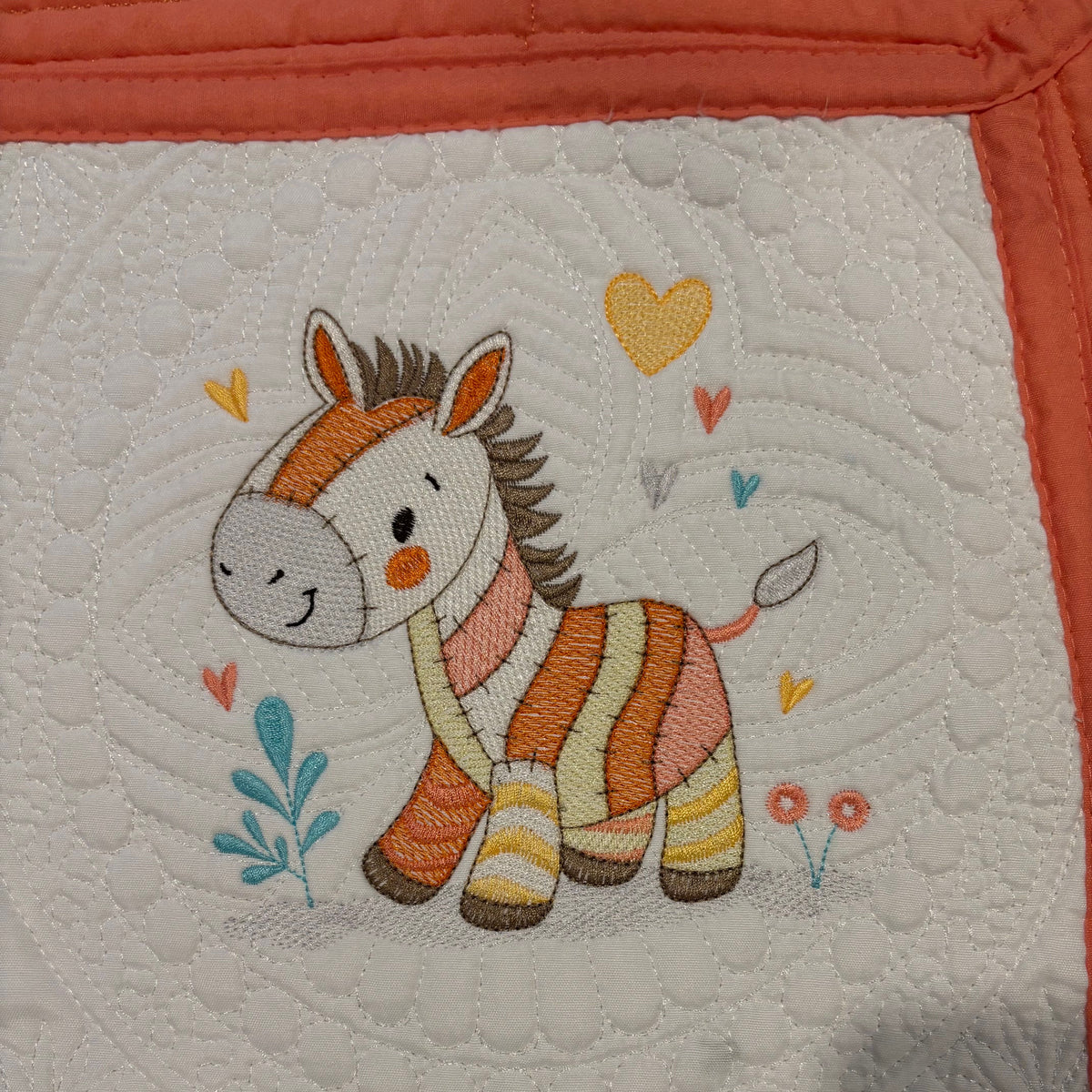 A close-up of a quilted fabric panel featuring an embroidered zebra with orange, cream, and brown stripes, surrounded by small embroidered hearts and leaves on a white background with a coral border.