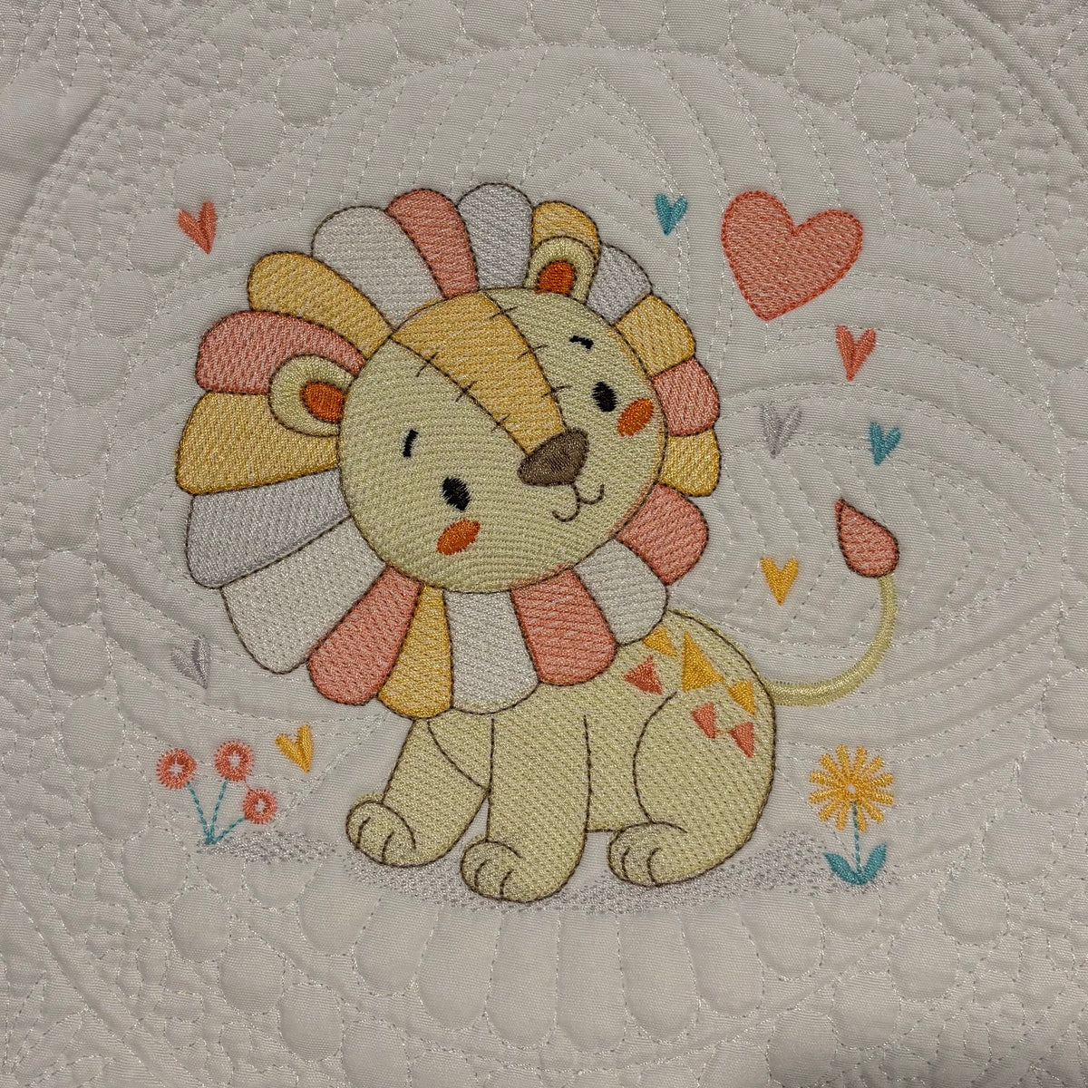 A close-up of a quilt featuring a colorful embroidered lion with a striped yellow, orange, and peach mane, surrounded by small embroidered hearts and flowers.
