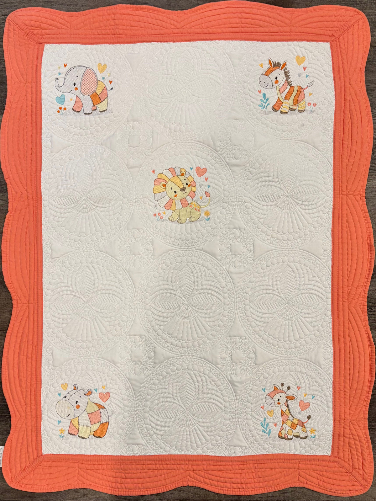 A baby quilt with a white center panel featuring five embroidered cartoon animals (elephant, hippo, lion, giraffe, zebra) surrounded by hearts, framed by a wide orange border on a dark wood background.