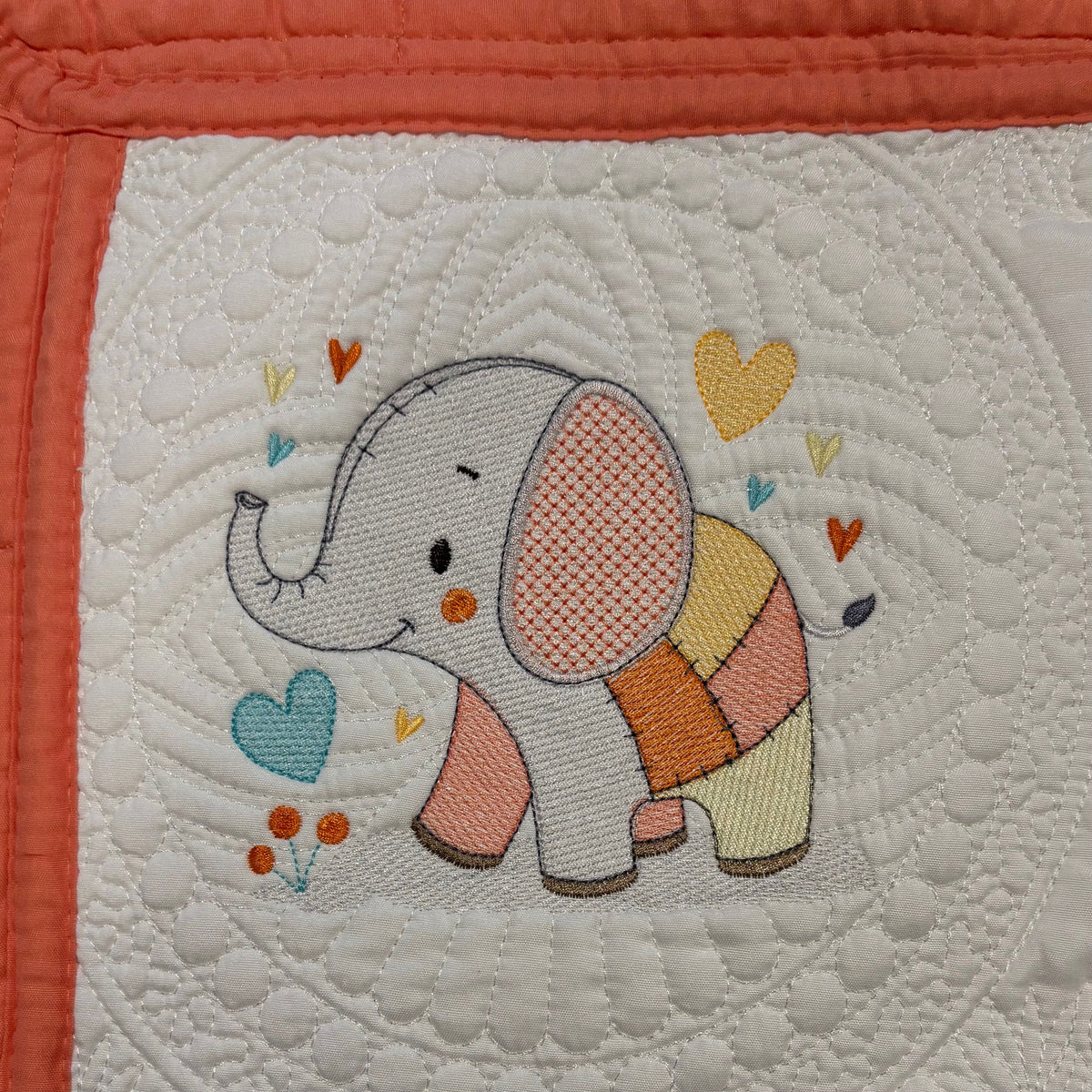 A close-up of a quilt featuring an embroidered elephant character with a peach patchwork body and colorful hearts and flowers, bordered by coral fabric.