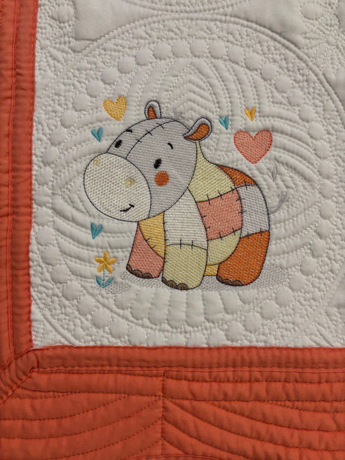 Close-up of a quilt featuring a stitched cartoon hippo in pastel colors, surrounded by small hearts and flowers, with an orange border and white quilted center.