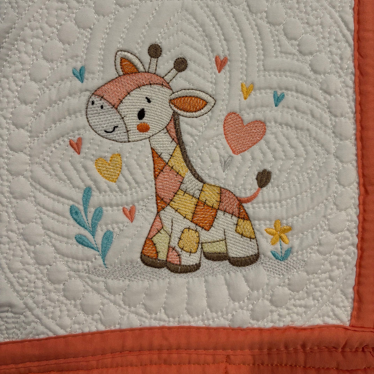 A close-up of a quilted fabric featuring a cartoon giraffe with a peach and orange checkered body, surrounded by embroidered hearts and leaves on a white background with an orange border.