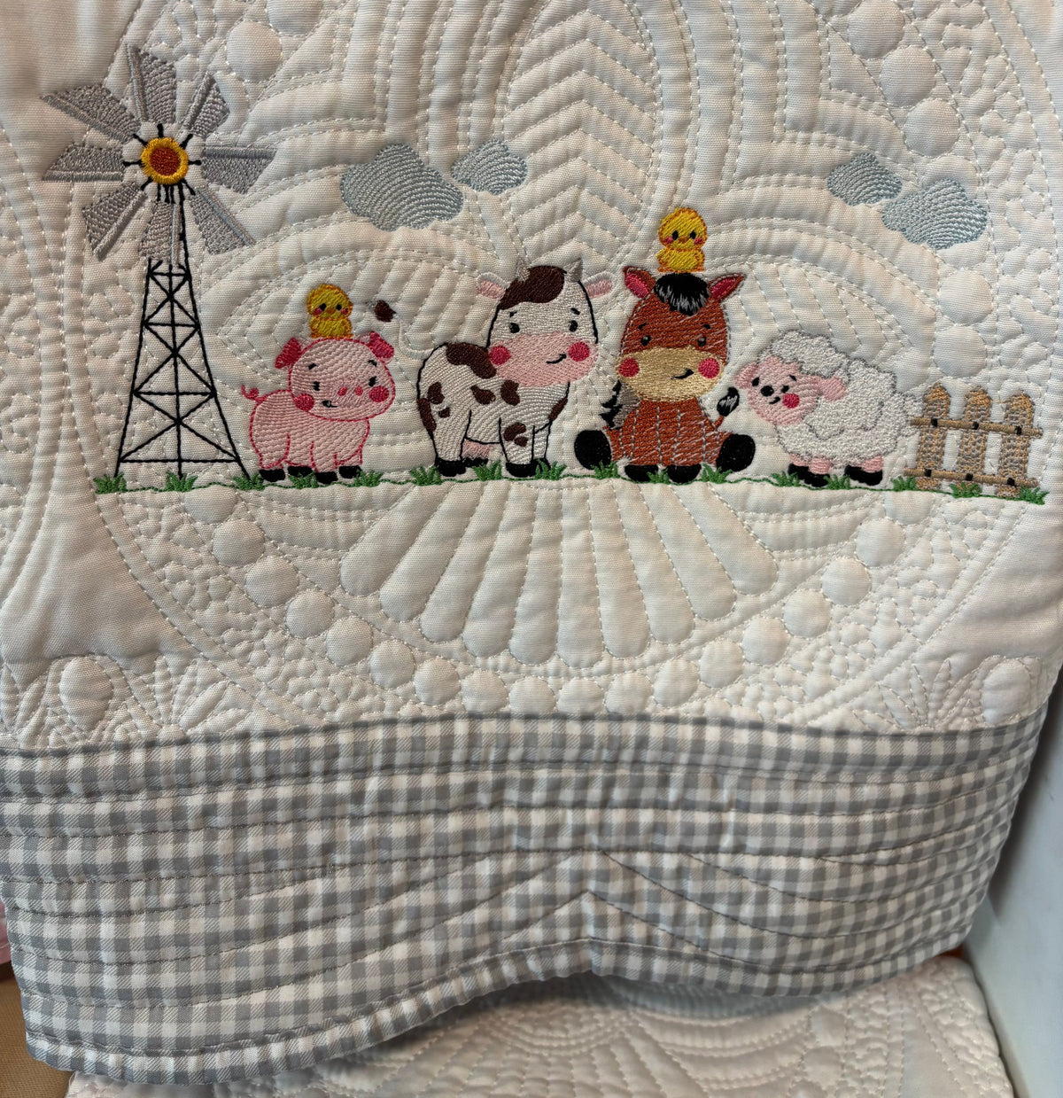 A close-up of a quilt featuring embroidered farm animals and a windmill, with a gray and white gingham check border along the bottom.