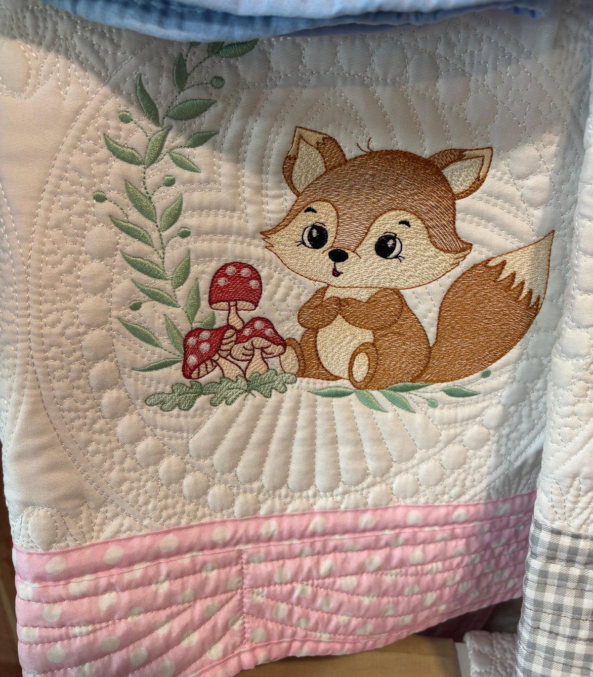 A close-up of a quilt featuring an embroidered brown fox, red-and-white mushrooms, green leaves, and a pink polka-dot stitched border.
