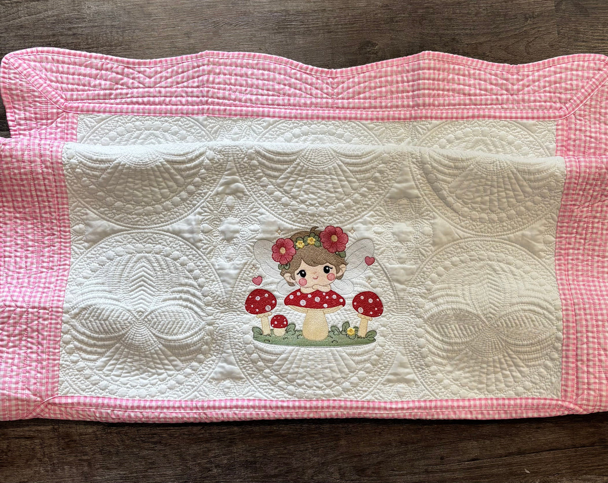 A pink and white gingham border on a baby quilt featuring a central embroidered design of a girl with flowers in her hair sitting on a red and white mushroom, displayed on a dark wood surface.