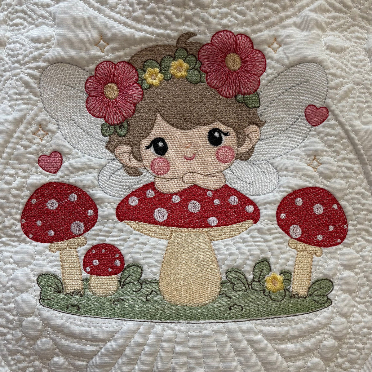 A close-up of a quilted fabric featuring an embroidered fairy design with red flowers in her hair, a mushroom cap under her, and red-and-white polka-dot mushrooms around her on a white quilted background.