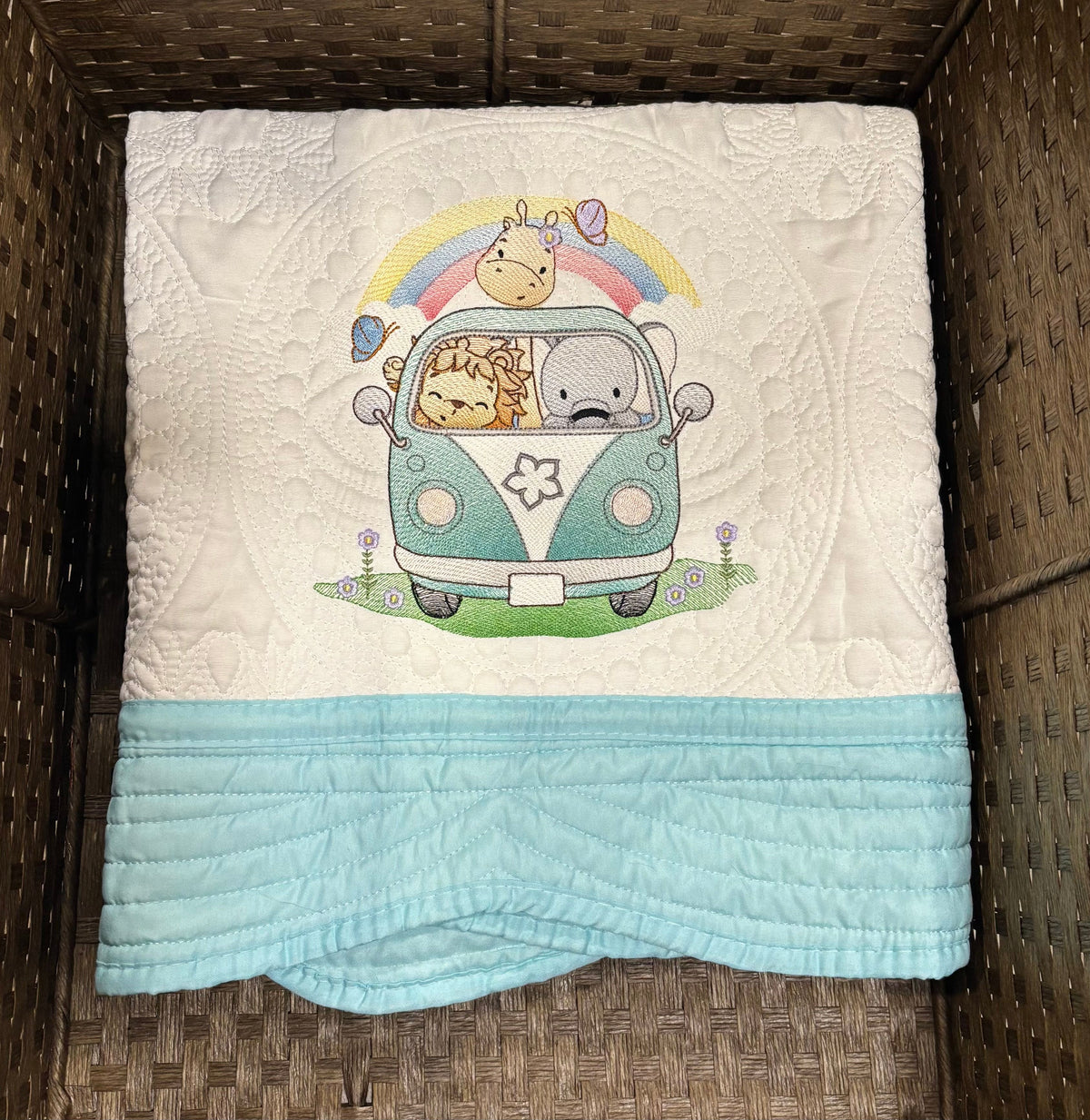 A baby quilt laid flat inside a brown wicker basket. The top section is white with a large embroidered illustration of a pastel rainbow, cute animals, and a mint green van.  The quilt has a pastel blue border.