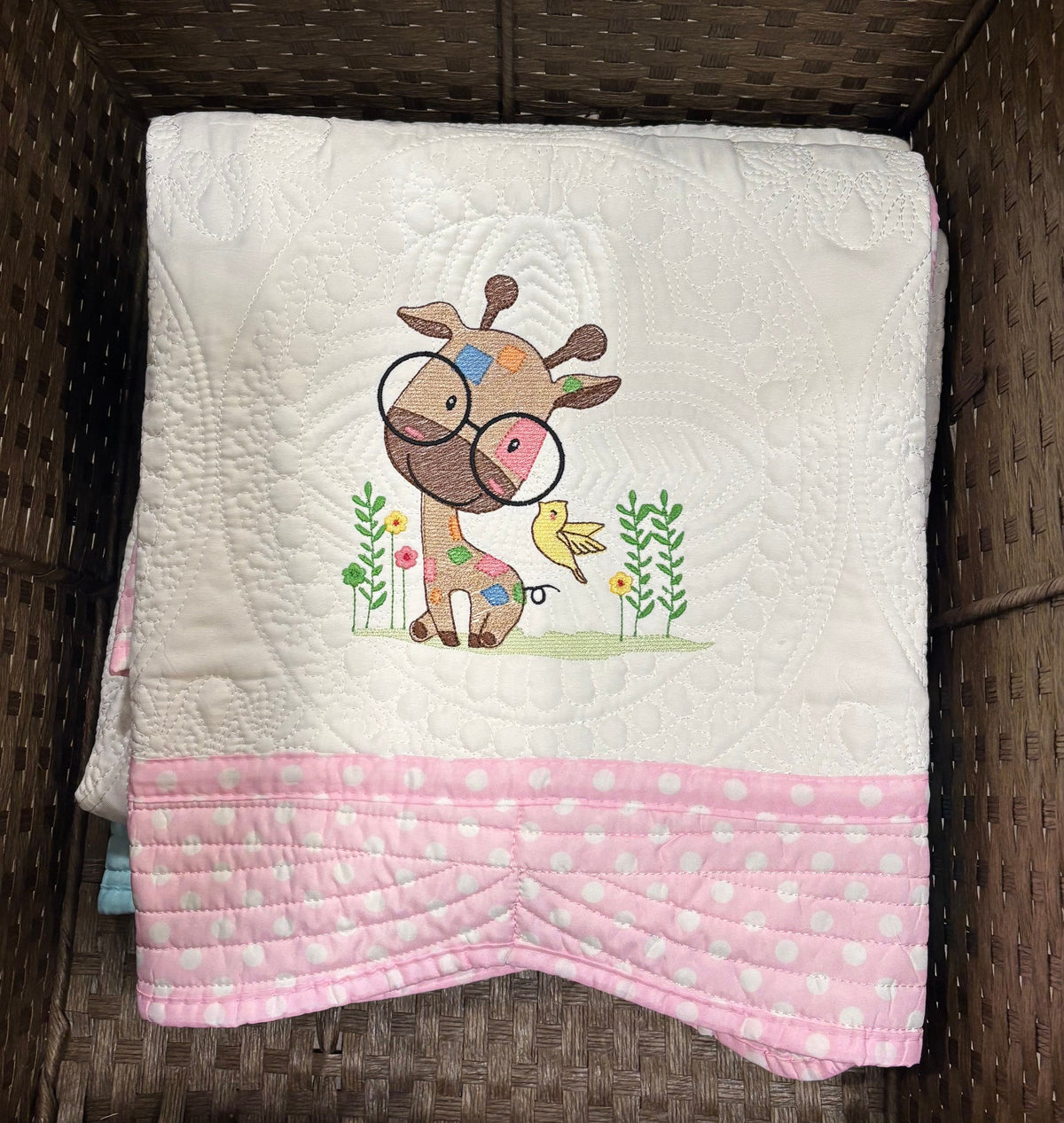 A folded white quilted baby blanket with a pink polka-dot hem and a central embroidered cartoon giraffe wearing glasses design, displayed inside a brown wicker basket.