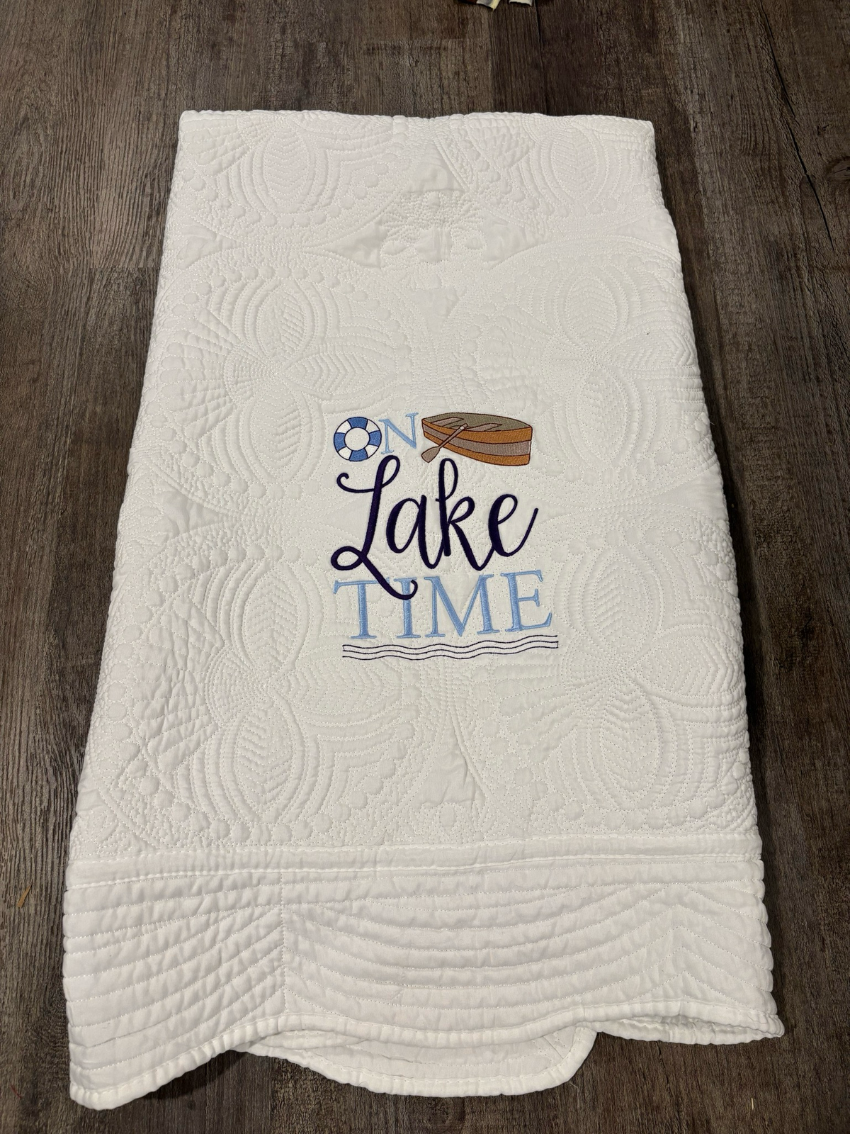 An off-white quilted cotton heirloom quilt with an 'On Lake Time' graphic, laid flat on a dark wood background. The design features navy and light blue lettering, a life ring, and a row boat.