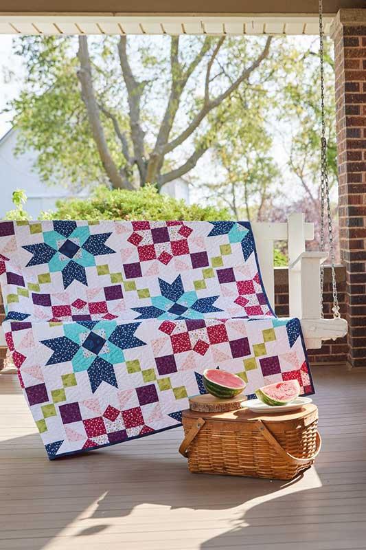 A colorful quilted throw blanket with a geometric star and checkered patch design is draped over a porch swing on a wooden porch. A wicker picnic basket with watermelons sits in front, with trees and a house visible in the background.