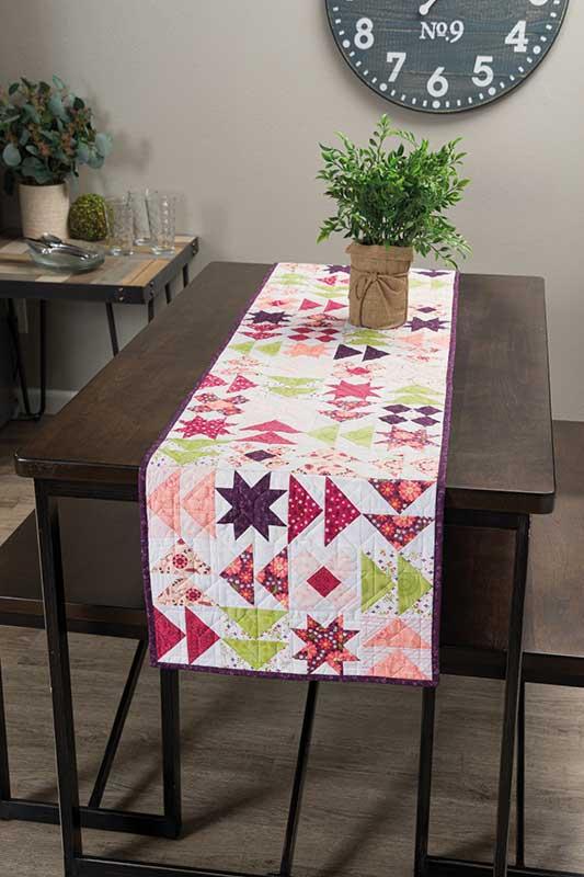 Quilted table runner on a wooden table with a clock and plants in the background.