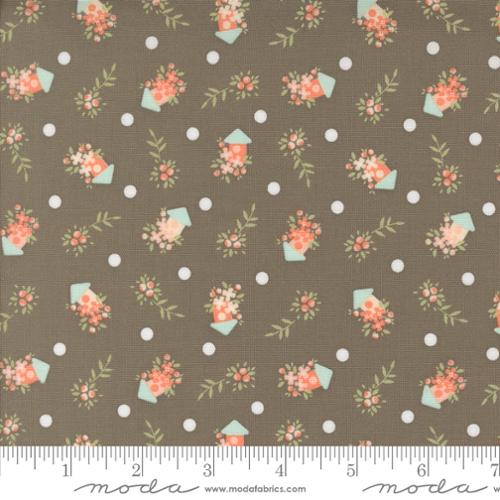 A close-up of a fabric pattern featuring a slate background with white polka dots, and small floral bouquets in coral and peach, sage green leaves, and mint green birdhouses. A white ruler is visible along the bottom edge, and the brand name 'moda' is printed on the ruler.