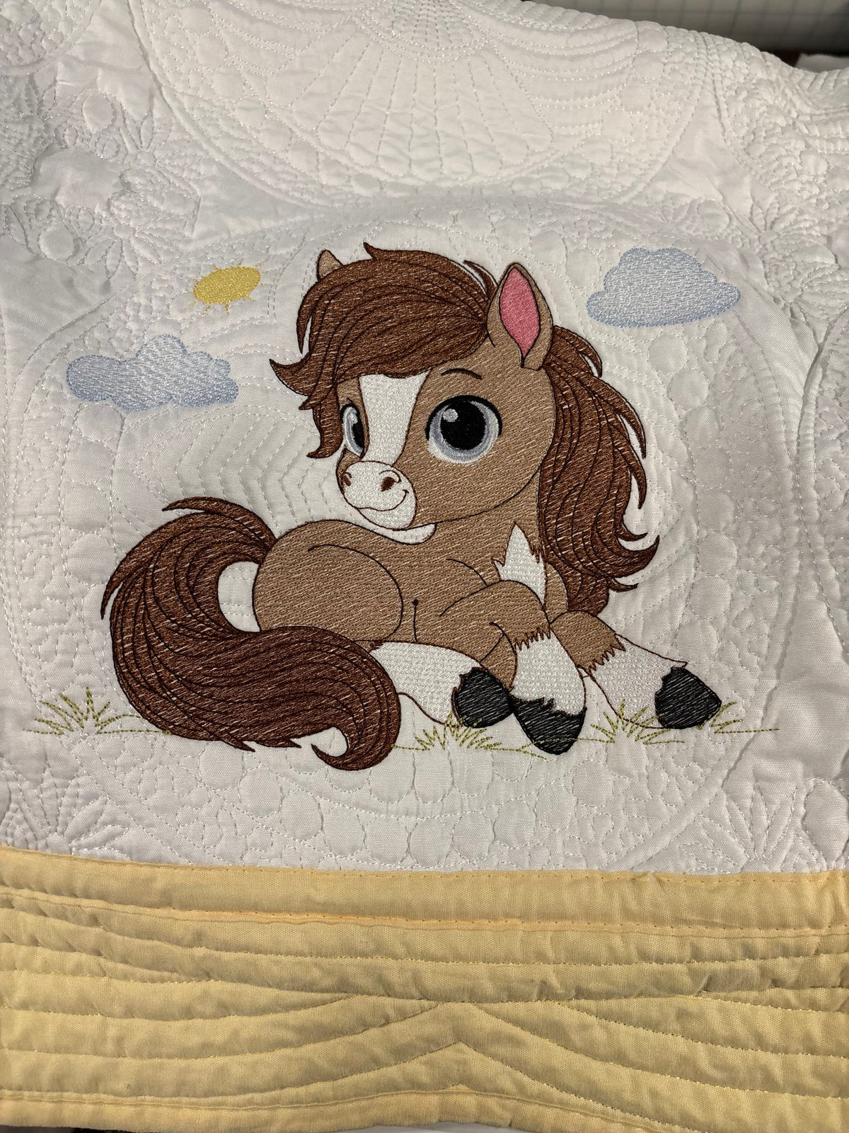 A close-up of a quilt featuring an embroidered cartoon brown horse with a white blaze, pink inner ear, and black hooves. A soft yellow quilted fabric band is stitched along the bottom edge.