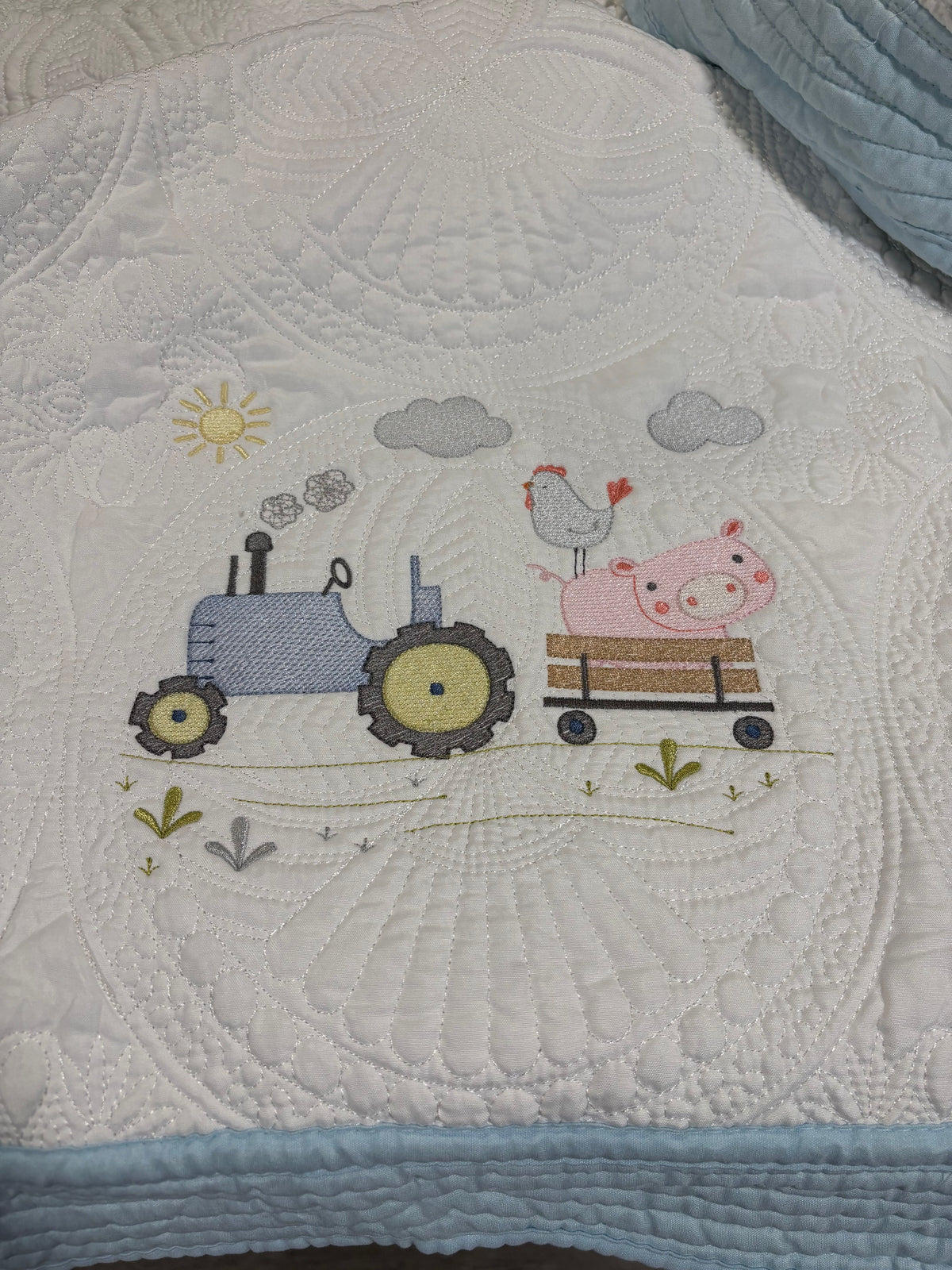 A close-up of a white quilted baby blanket featuring a colorful embroidered design of a blue tractor, a pink pig on a wooden cart, a chicken, clouds, and a sun, finished with a light blue border.