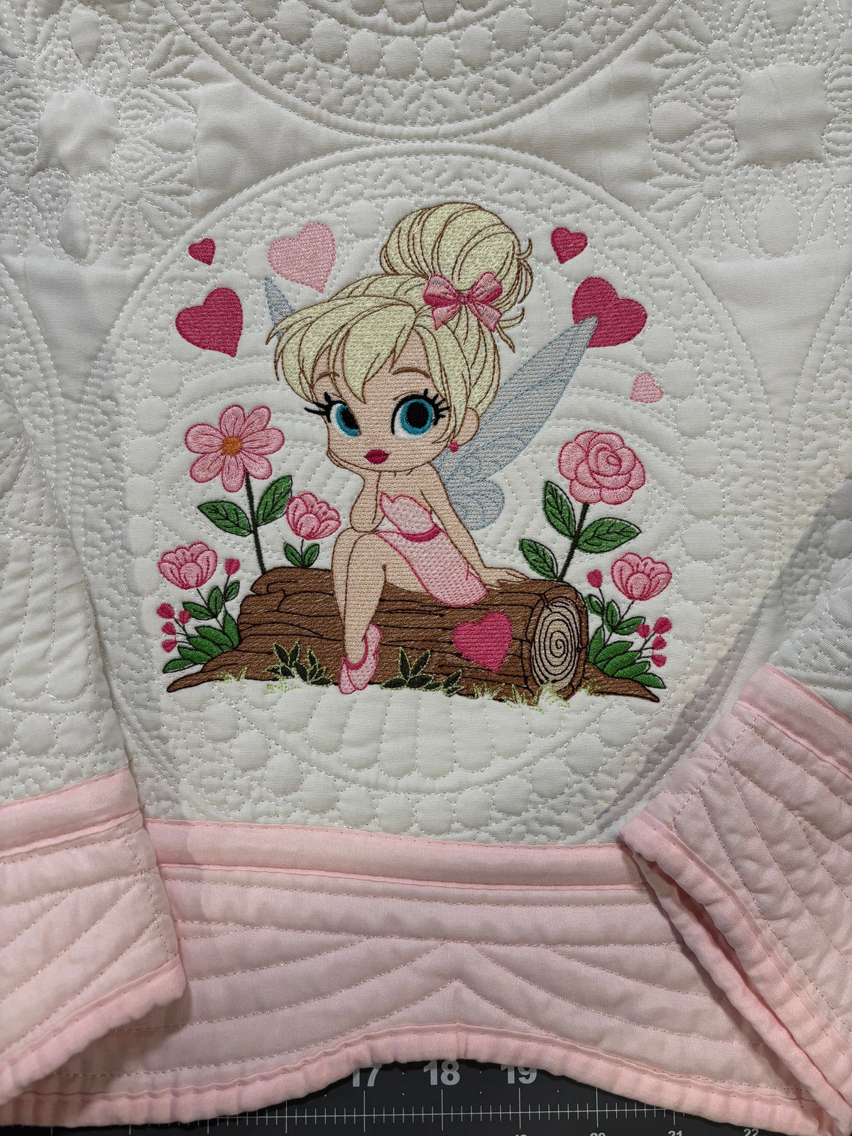A close-up of a quilt featuring an embroidered fairy design. The fairy has blonde hair in a bun, blue eyes, pink wings, and is sitting on a log with pink flowers and hearts around her.