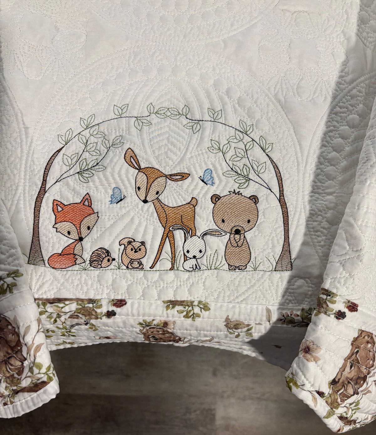 A close-up of a white quilt featuring a woodland animal design with a fox, deer, bear, rabbit, squirrel and small hedgehog under a tree arch.  There are also 2 blue butterflies in the design. The quilt is draped over a bed or chair, with a gray-brown floor visible underneath.