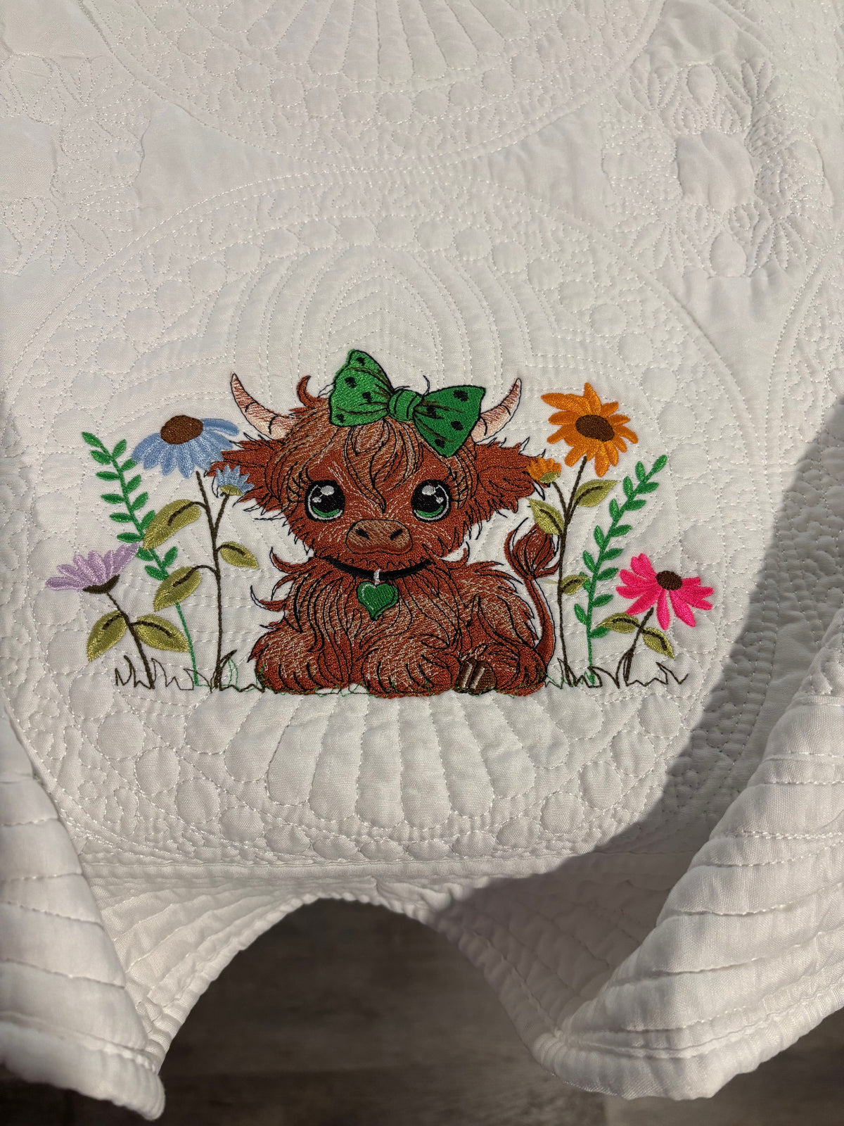 A close-up of a white quilted blanket, featuring an embroidered Scottish highland cow with a green bow and colorful wildflowers.