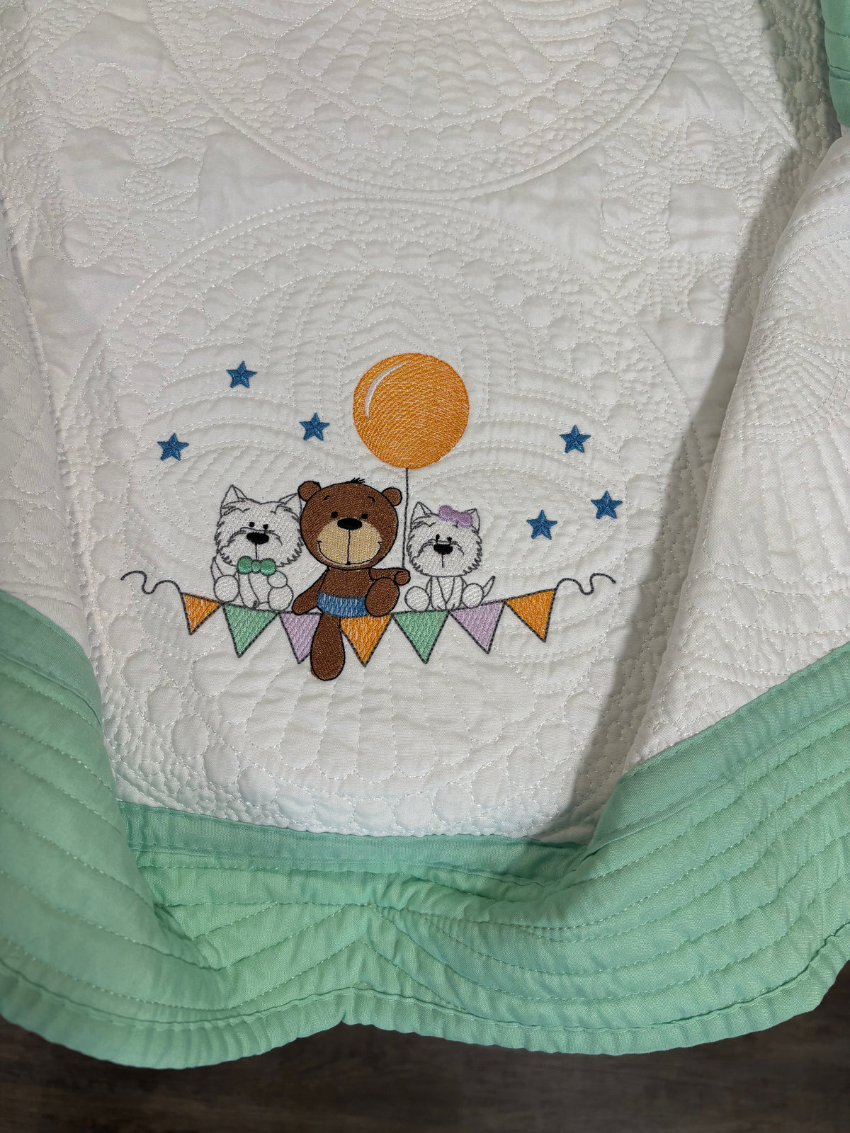 A close-up of a white quilt with mint green trim, featuring an embroidered design of a brown teddy bear, two white dogs, blue stars, and an orange balloon.