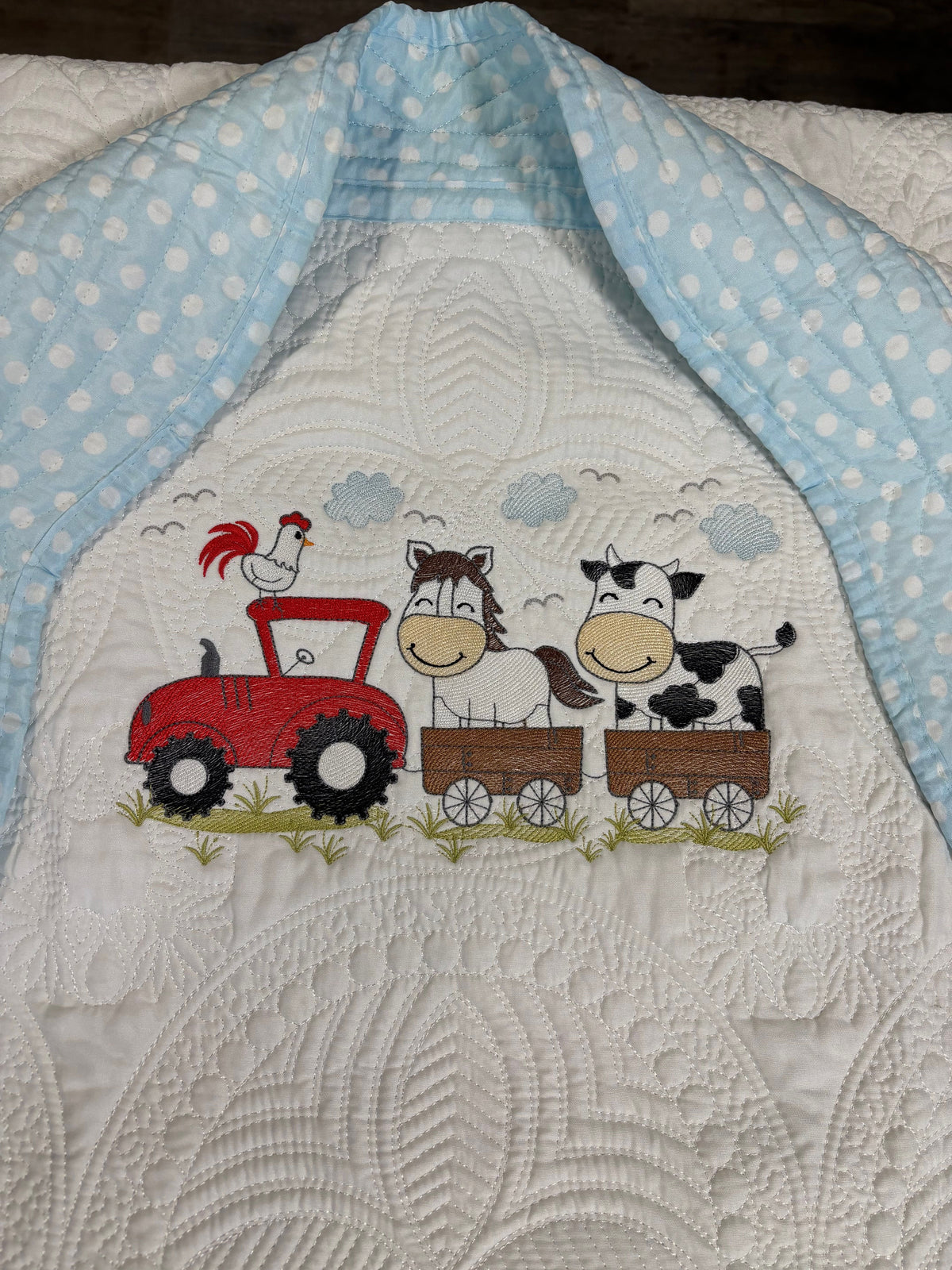A close-up of a quilt with a farm-themed embroidery design. The white quilted fabric features a red tractor carrying a brown horse in a wagon and a black-and-white cow in another wagon, with a red rooster in a red tractor and small blue clouds above.