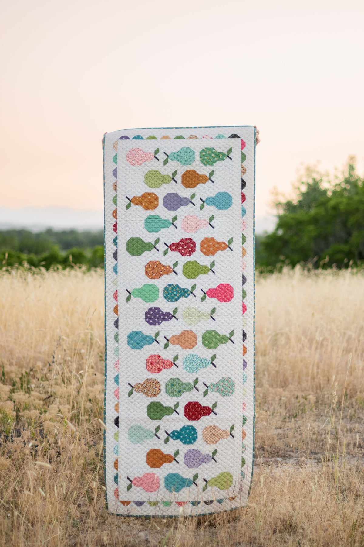 A rectangular quilted table runner with a colorful pattern of stylized pears, standing upright in a field of dry grass at sunset.
