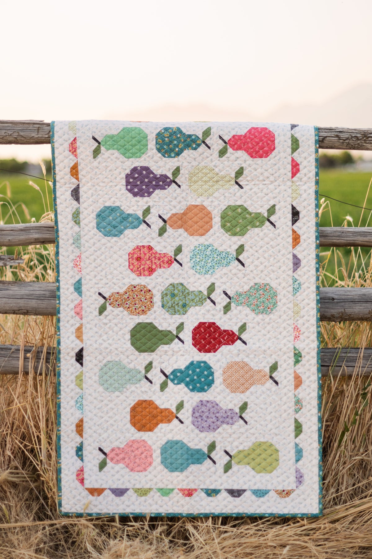 A quilted table runner with a colorful pears pattern draped over a rustic wooden fence in a field.