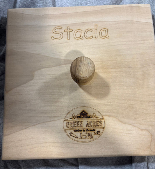 A personalized light wood quilting clapper with a round wooden knob, engraved with a name at the top and a circular 'Green Acres' logo at the bottom, shown on a gray fabric background.