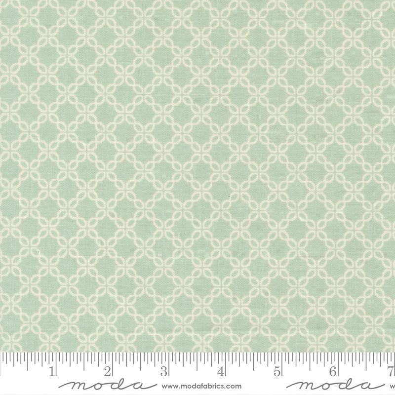 A close-up of a fabric pattern with a pale blue background and a repeating cream-colored geometric trellis design. A white fabric ruler is visible along the bottom edge to show scale, with the Moda brand name and website printed on the ruler.