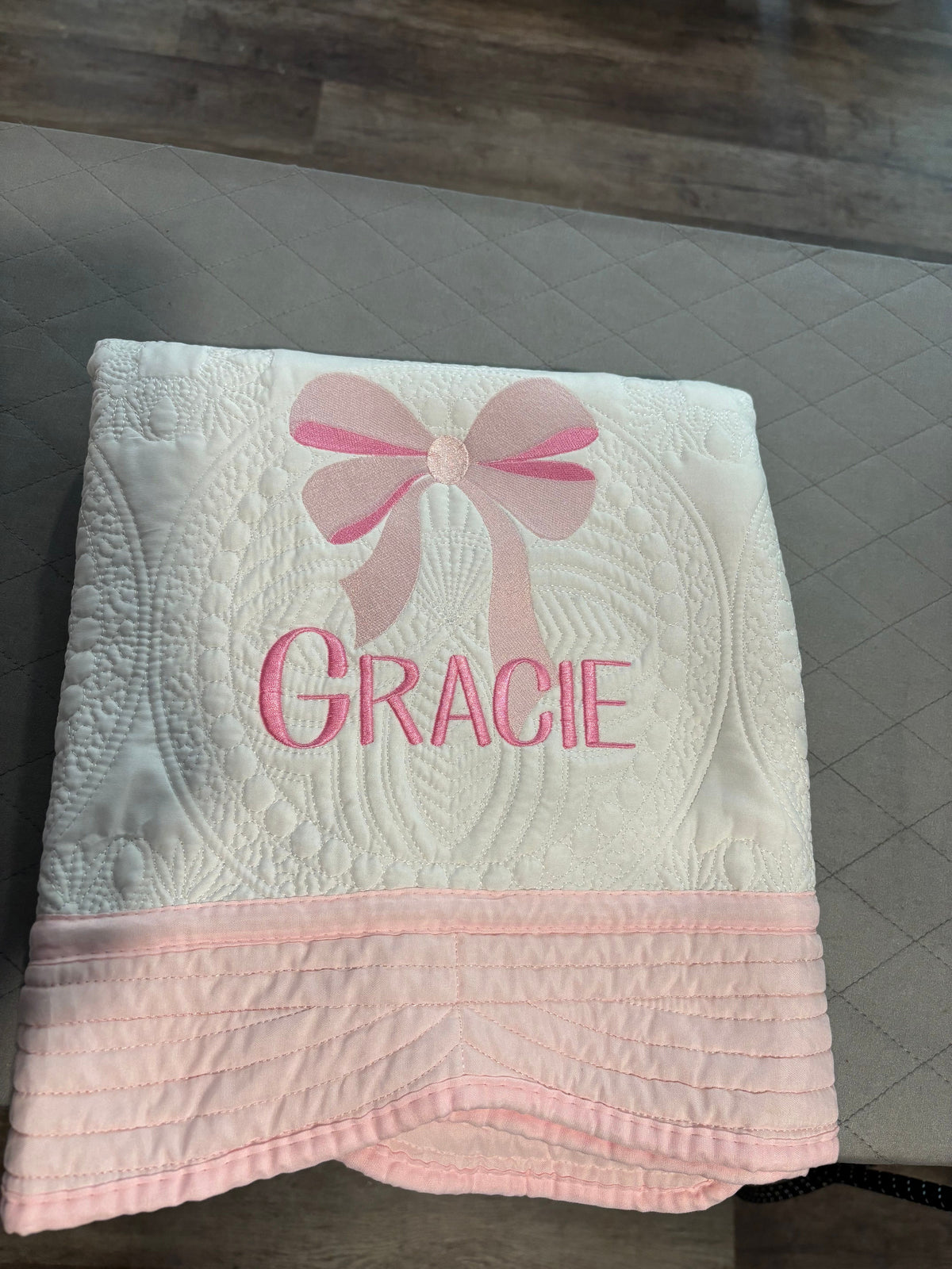 A personalized baby blanket with a white quilted top section, a pink bow design, and the name 'GRACIE' embroidered in pink. The bottom of the blanket features a solid pink border.