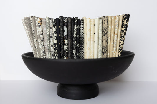 A black pedestal bowl filled with a vertical row of folded fabric swatches in black, white, beige, and gray patterns, set against a plain white background.