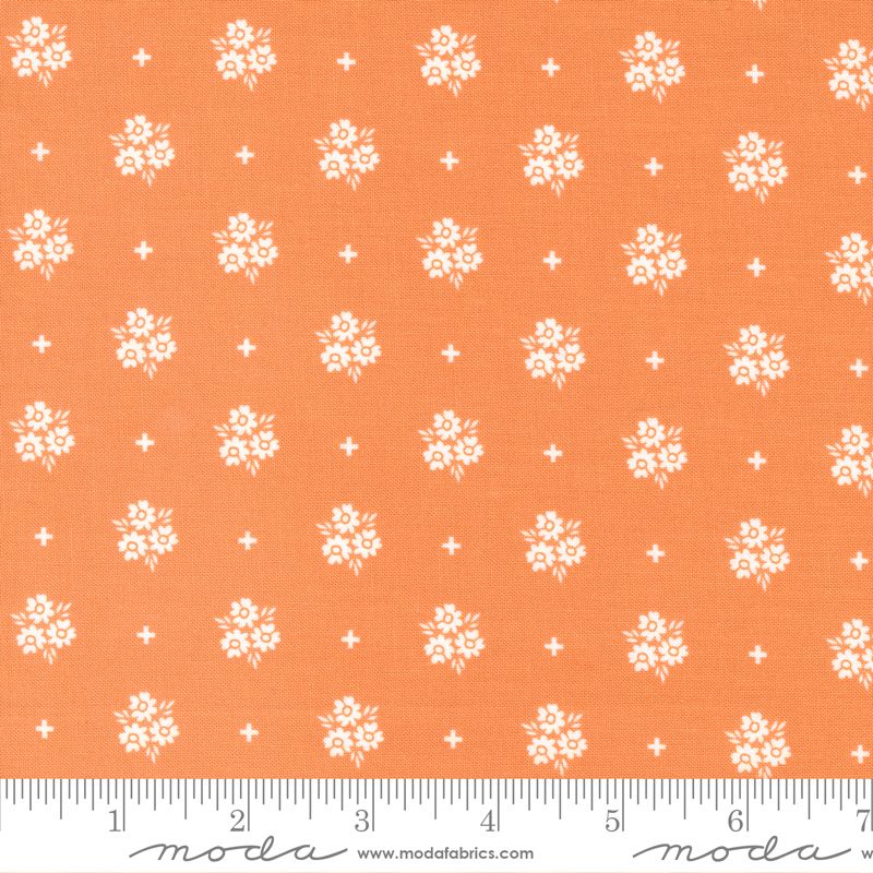 A close-up of a fabric pattern featuring white floral motifs and small white plus signs on a warm orange background, with a white ruler along the bottom edge showing measurements. The brand name 'Moda' is visible on the ruler watermark.