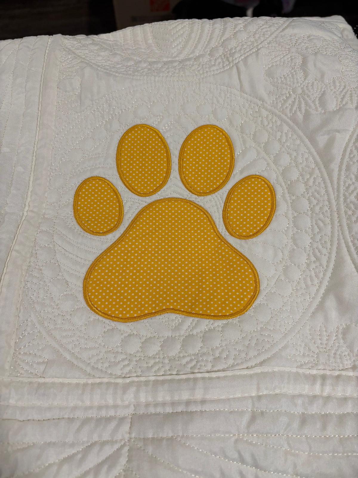 A close-up of a white quilted blanket featuring a large yellow paw print applique with a dotted texture.