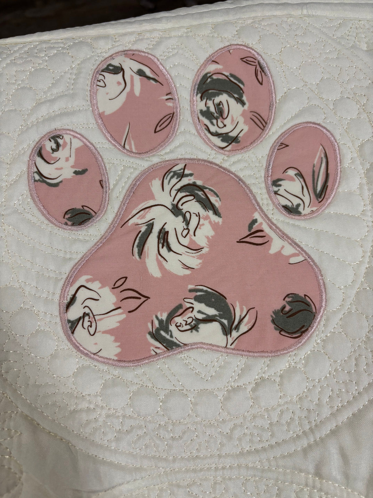 A close-up of a quilted white fabric item featuring a large pink paw print applique with a floral pattern.
