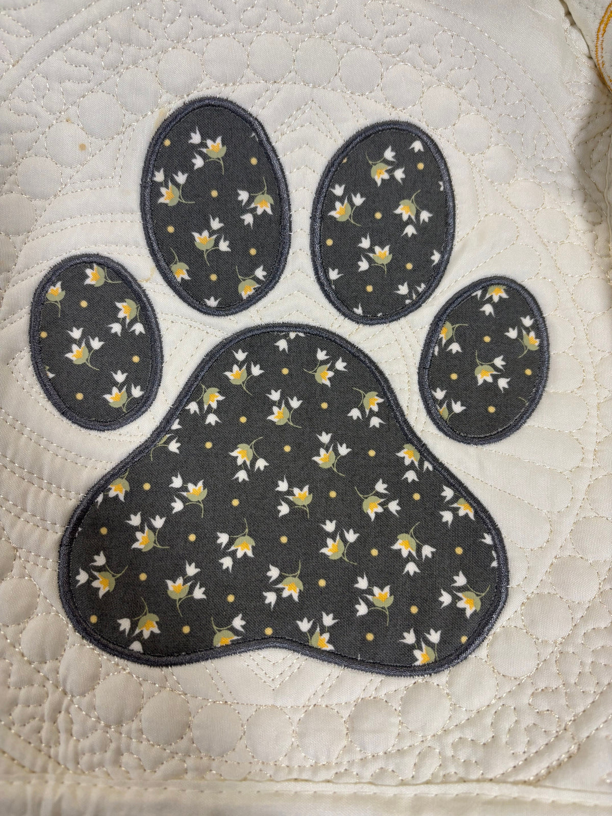 A quilted fabric item featuring a large paw print applique in dark gray with a white and yellow floral pattern, set against a cream-colored quilted background.