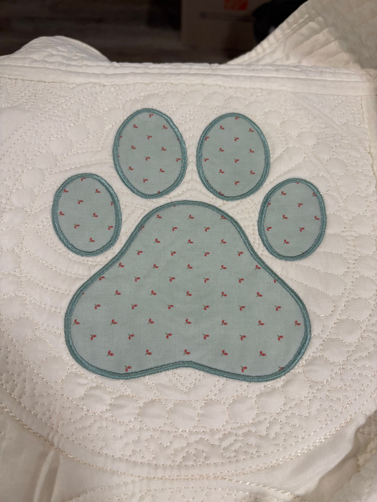 A close-up of a white quilt featuring a large embroidered or applique paw print in a pale blue-green color with tiny red embroidered details.