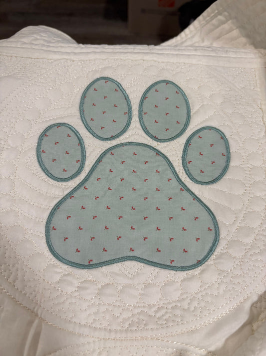 A close-up of a white quilt featuring a large embroidered or applique paw print in a pale blue-green color with tiny red embroidered details.